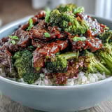 A close-up of a homemade Beef and Broccoli Bowl shows glossy sauce drizzling over rice, beef, and bright green broccoli florets.